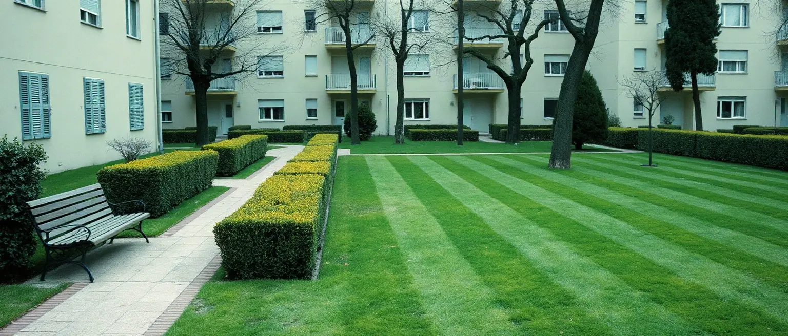Il verde comune di un condominio con il prato tagliato a strisce, le siepi squadrate e una panchina lungo il vialetto.